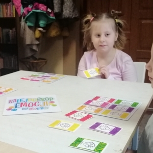 Przy stole, na którym leża kolorowe karty siedzą trzy dziewczynki.