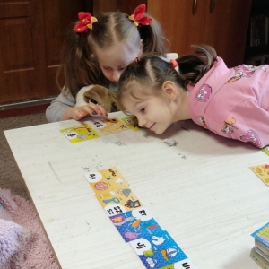 Dziecko połową ciała leży na stole, dwie pozostałe dzie2wczynki siedzą przy stole. Na stole rozłożone są karty do gry.