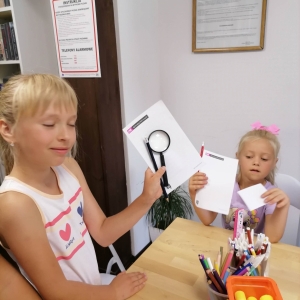 Dziewczynki siedząc przy stole trzymając kartki, ołówki i lupę